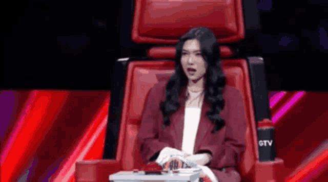 a woman in a red jacket is sitting in a red chair on a stage with her mouth open .