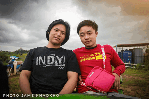 a man wearing a black indy shirt sits next to another man in a red supreme shirt