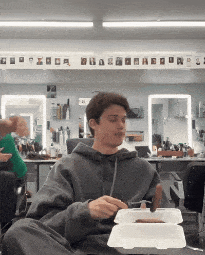 a man in a grey hoodie is eating a hot dog from a styrofoam box
