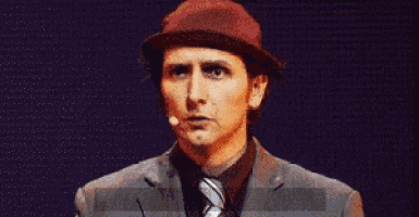 a man wearing a hat and a suit says what the fuck in front of a microphone .