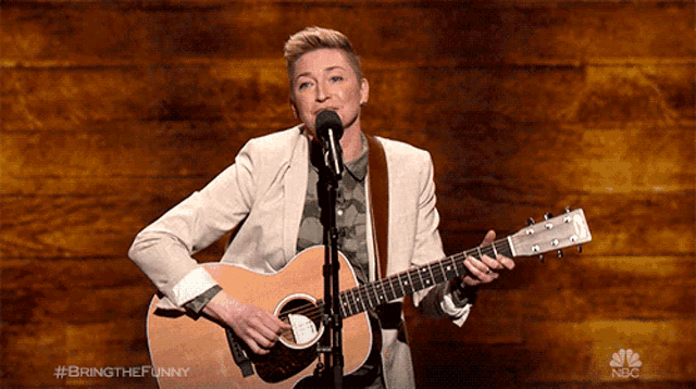 a woman is singing into a microphone while holding a guitar in front of a nbc logo