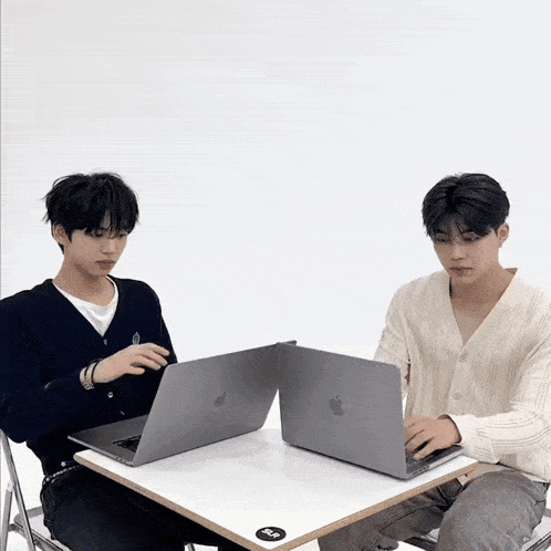 two young men are sitting at a table with laptops and one of them is wearing a white sweater