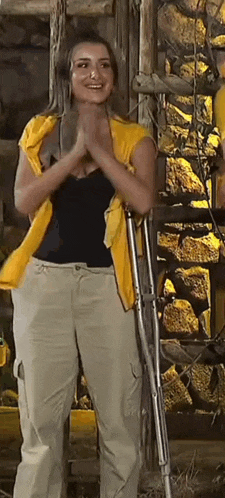 a woman wearing a yellow vest and khaki pants is standing in front of a stone wall with crutches .