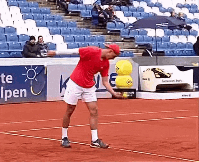 a man in a red shirt stands on a tennis court in front of a sign that says te alpen