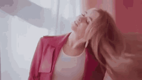 a woman in a pink jacket is standing in front of a window with her eyes closed and her hair blowing in the wind .