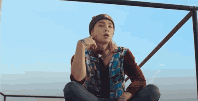 a young man wearing a beanie and a plaid shirt is sitting on a railing looking at the camera .