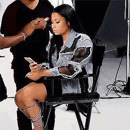 a woman in a denim jacket is sitting in a chair looking at her cell phone .