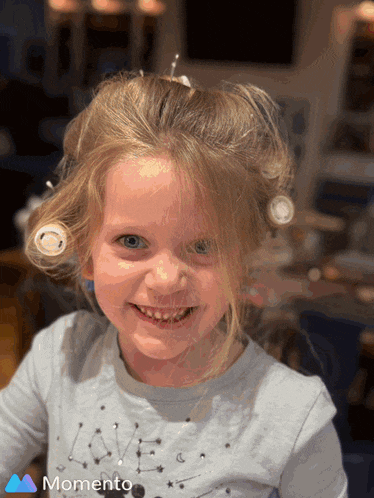 a little girl with curlers in her hair is smiling and wearing a momento shirt