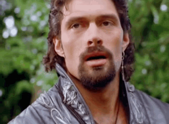 a man with a beard and long hair is wearing a leather jacket and earrings .