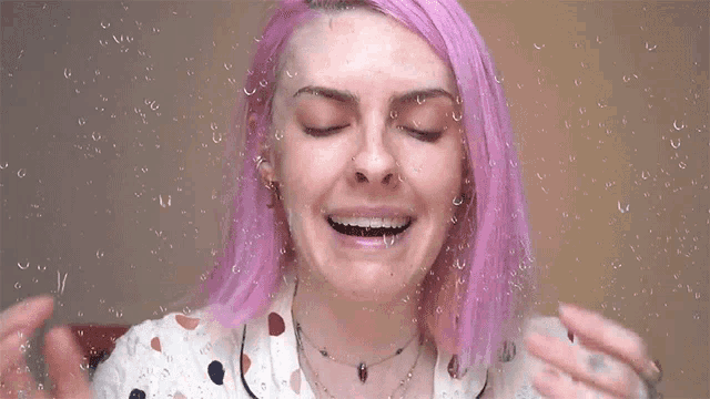 a woman with pink hair is smiling in front of a glass with water drops on it