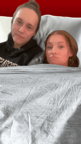 two women are laying on a bed with one wearing a hoodie that says ' nc state ' on the front