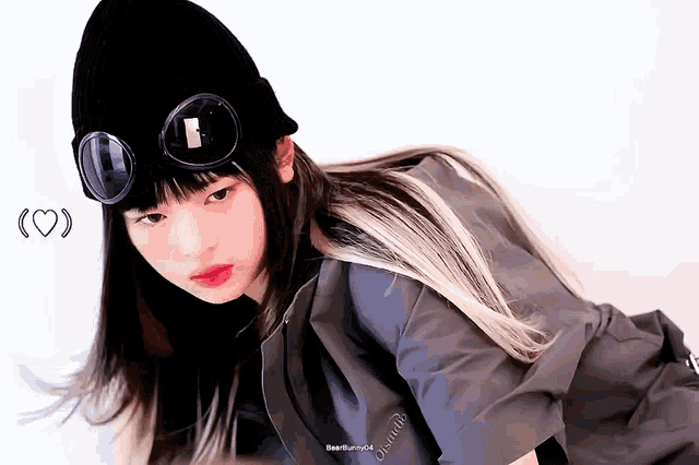 a girl wearing a black beanie and goggles looks at the camera with a heart behind her