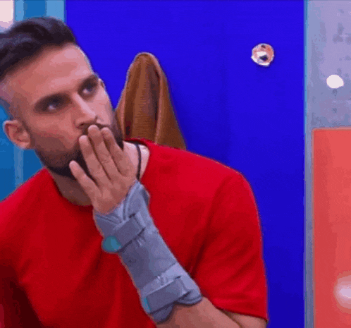 a man in a red shirt with a cast on his arm is covering his mouth with his hand .