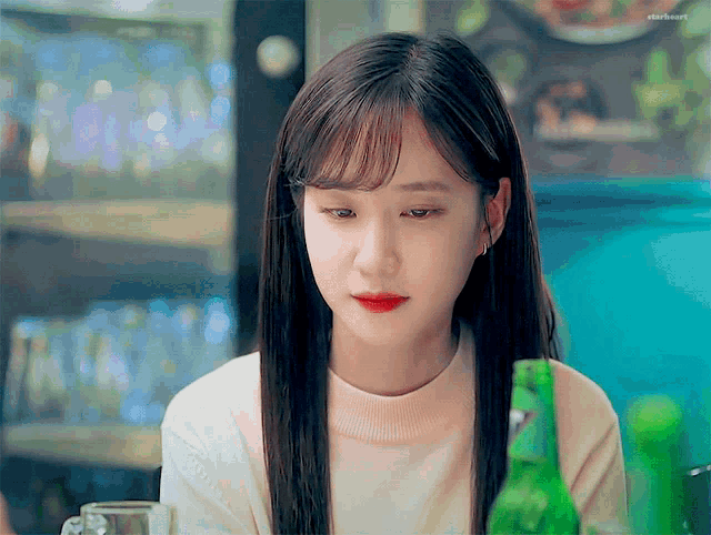 a woman with long hair is sitting at a table with a bottle of beer in front of her