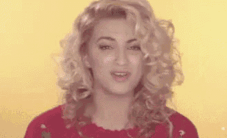 a woman with curly blonde hair is wearing a red sweater and making a funny face .