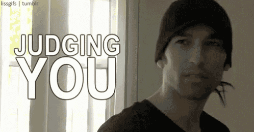 a man wearing a black beanie is standing in front of a window with the words judging you above him