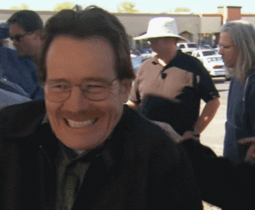 a man wearing glasses and a hat smiles in front of a group of people