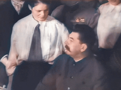 a man with a mustache is sitting in a crowd of people and talking to another man .