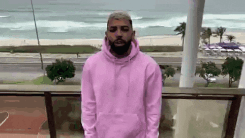 a man with a beard is wearing a pink hoodie and standing on a balcony .
