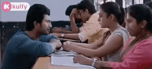 a man and a woman are sitting at a table in a classroom with other people .