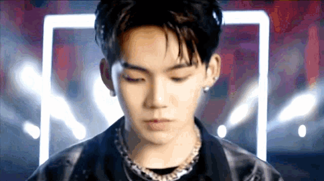 a young man wearing a black jacket and a necklace is looking down .