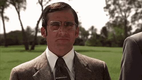 a man in a suit and tie is standing in a grassy field with trees in the background .