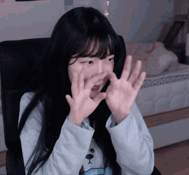 a girl with long black hair is sitting in a chair and covering her mouth with her hands
