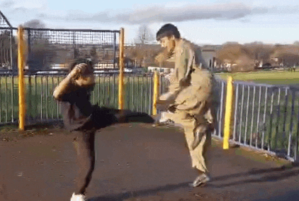 a man is kicking another man in the face while standing next to a fence in a park .