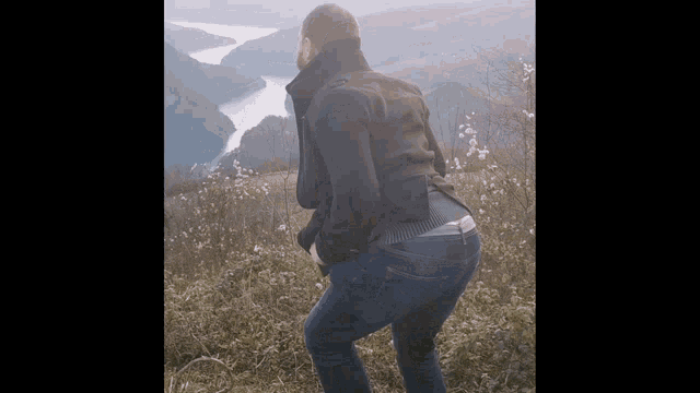 a man in a black jacket and blue jeans is standing on a hill overlooking a river