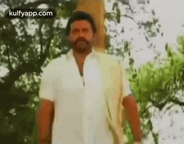 a man with a beard and a white shirt is standing in front of trees .