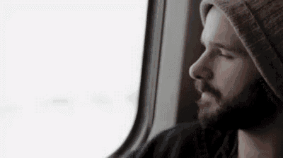 a man with a beard is looking out of a train window while wearing a hat .