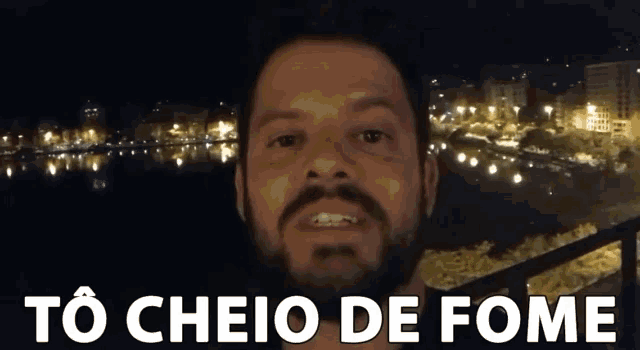 a man with a beard stands in front of a city at night with the words to cheio de fome below him