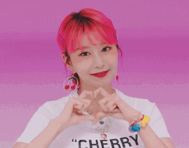 a girl with pink hair wearing a cherry shirt makes a heart with her hands
