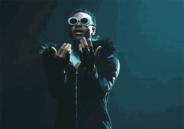 a man wearing sunglasses and a black jacket holds his hands in the air