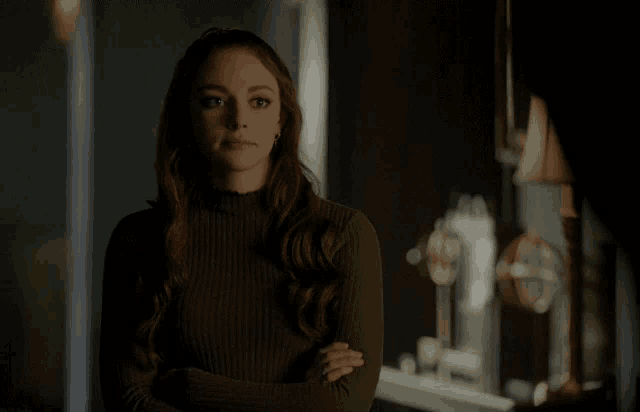 a woman in a green sweater stands with her arms crossed in a dark room