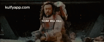 a man with a beard is holding a gun in front of a fire and a sign that says `` some will fall '' .