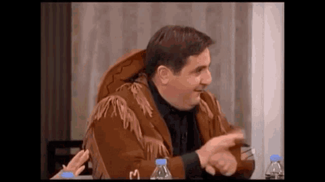 a man in a fringed jacket is sitting at a table with bottles of water .