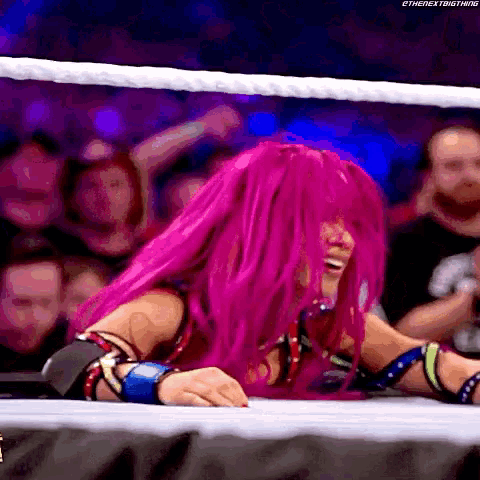 a woman with pink hair is sitting in a wrestling ring with a crowd behind her .
