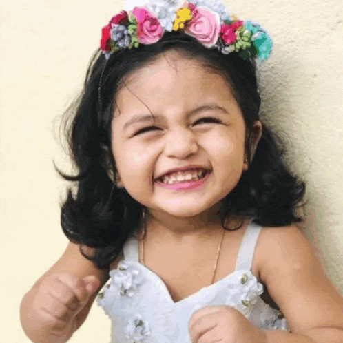 a little girl wearing a flower crown and a white dress is smiling for the camera .