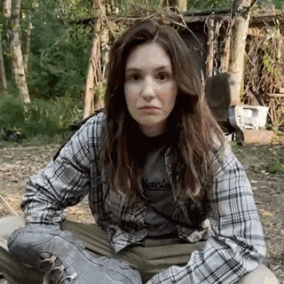 a young woman in a plaid shirt is sitting on the ground in the woods .