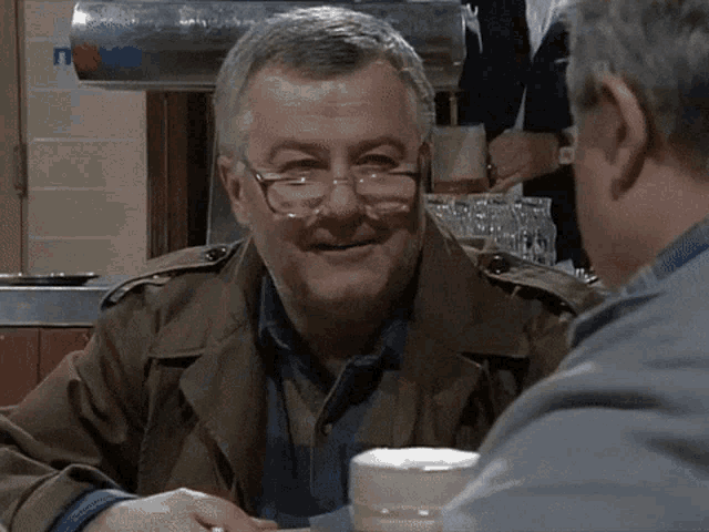a man wearing glasses sits at a table with another man in front of a tv that says nova