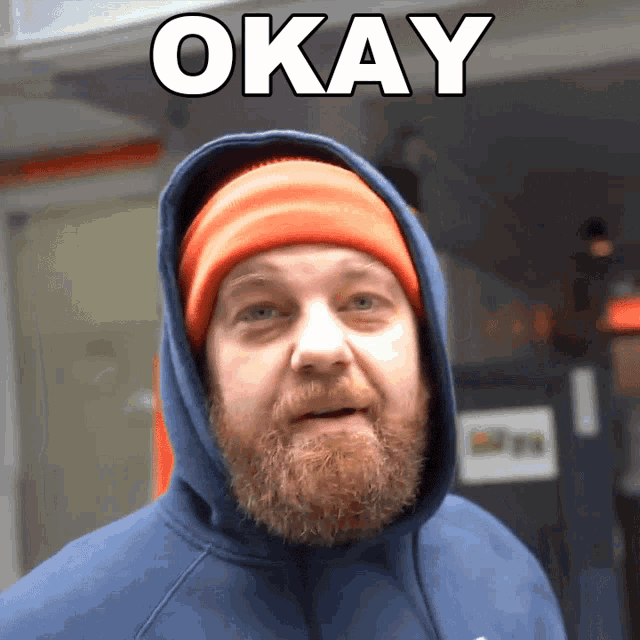a man with a beard wearing a blue hoodie and an orange beanie has the word okay above his head