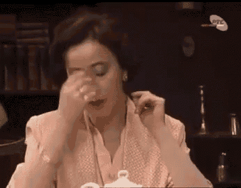 a woman is sitting at a table with a glass of water in her hand and rubbing her eyes .
