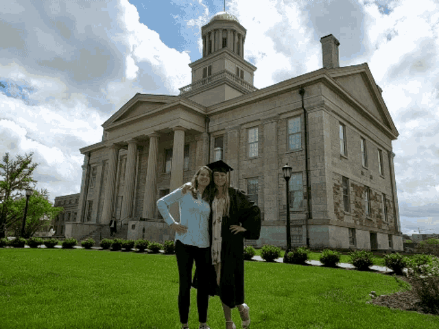 a woman in a graduation cap and gown poses with another woman in front of a large building