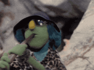 a green puppet wearing sunglasses and a hat is holding a stick