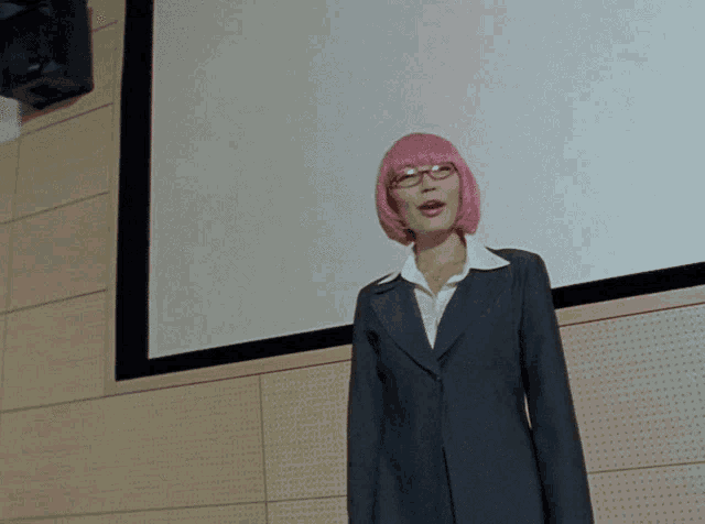 a woman with pink hair and glasses is standing in front of a white screen