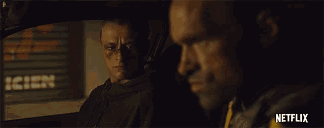 two men are looking at each other in a car with netflix written on the bottom right