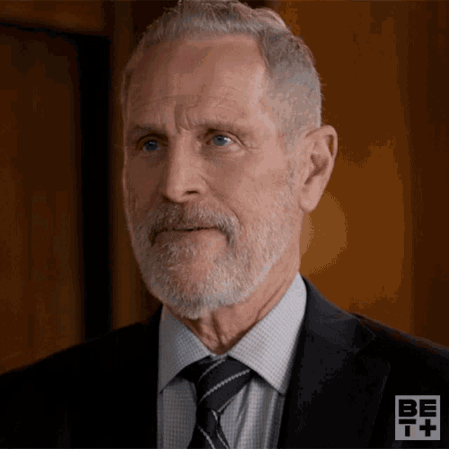 a man with a beard is wearing a suit and tie with a be t + logo in the background