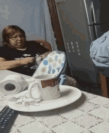 a woman sits at a table with a cup and a remote control on it