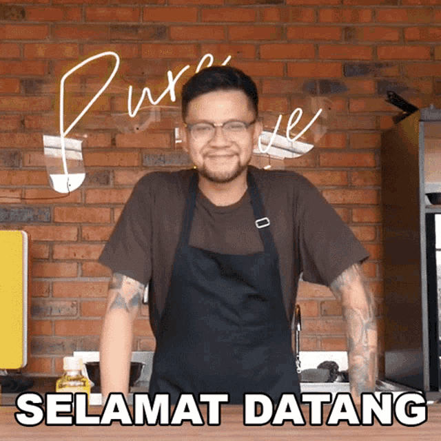 a man wearing an apron and glasses stands in front of a brick wall and says selamat datang